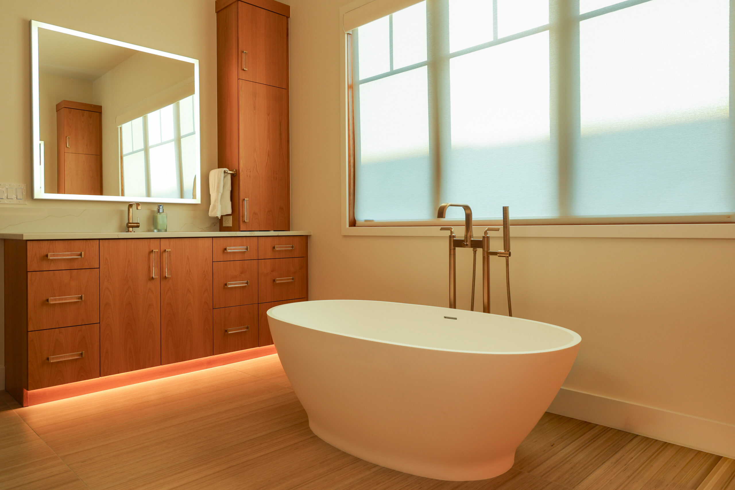 A brightly lit bathroom with a stand alone bathtub and light wood colored cabinets.
