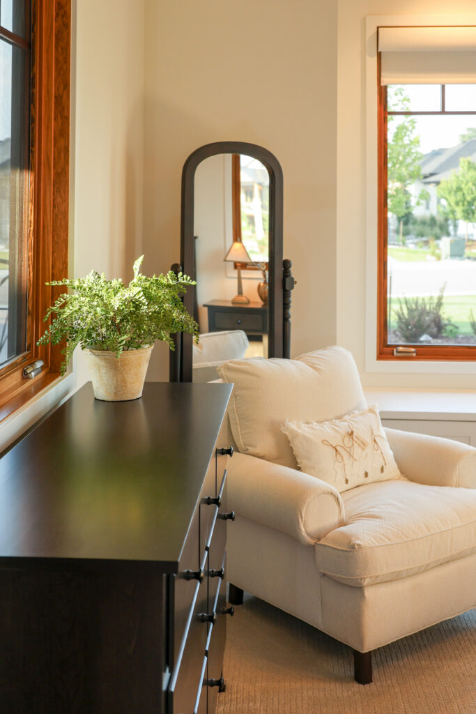 The corner of a bedroom with a black dresser, black standing mirror, white armchair, and windows.