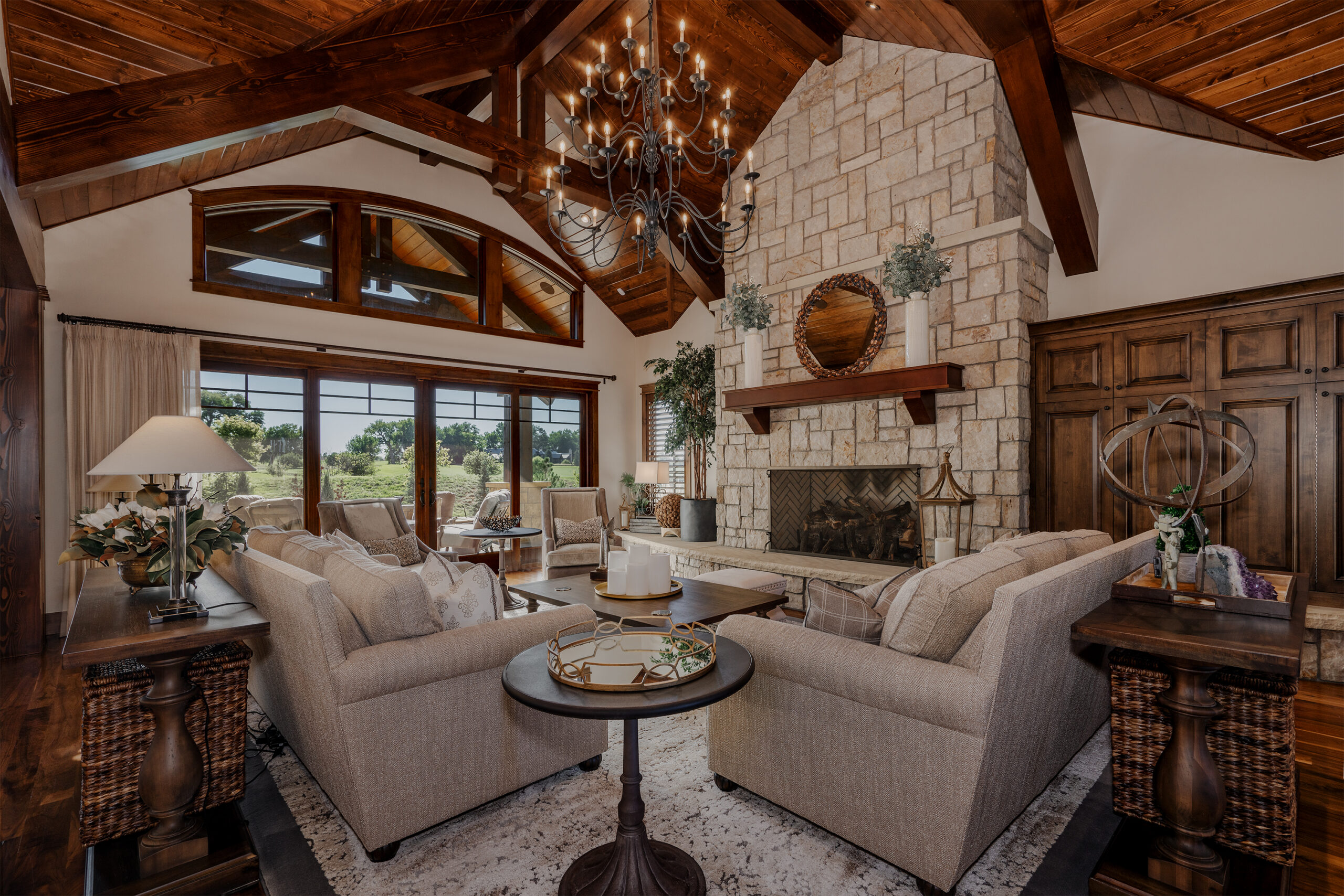 A large living room with a bright chandelier, large windows, a floor to ceiling stone fireplace, and sofa and chairs.