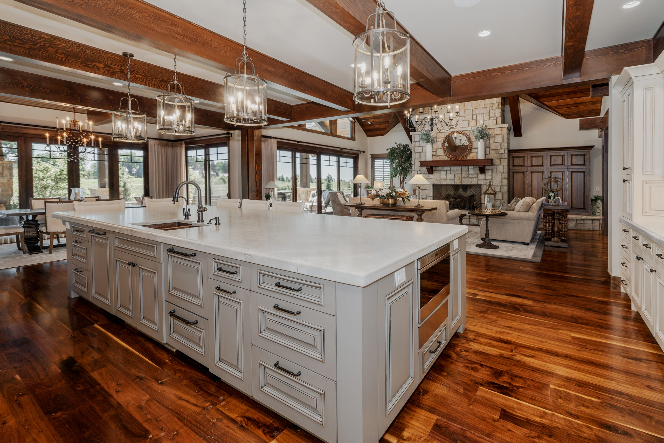 A kitchen with a large island and hanging lights looking into a living room space with sofas and windows.