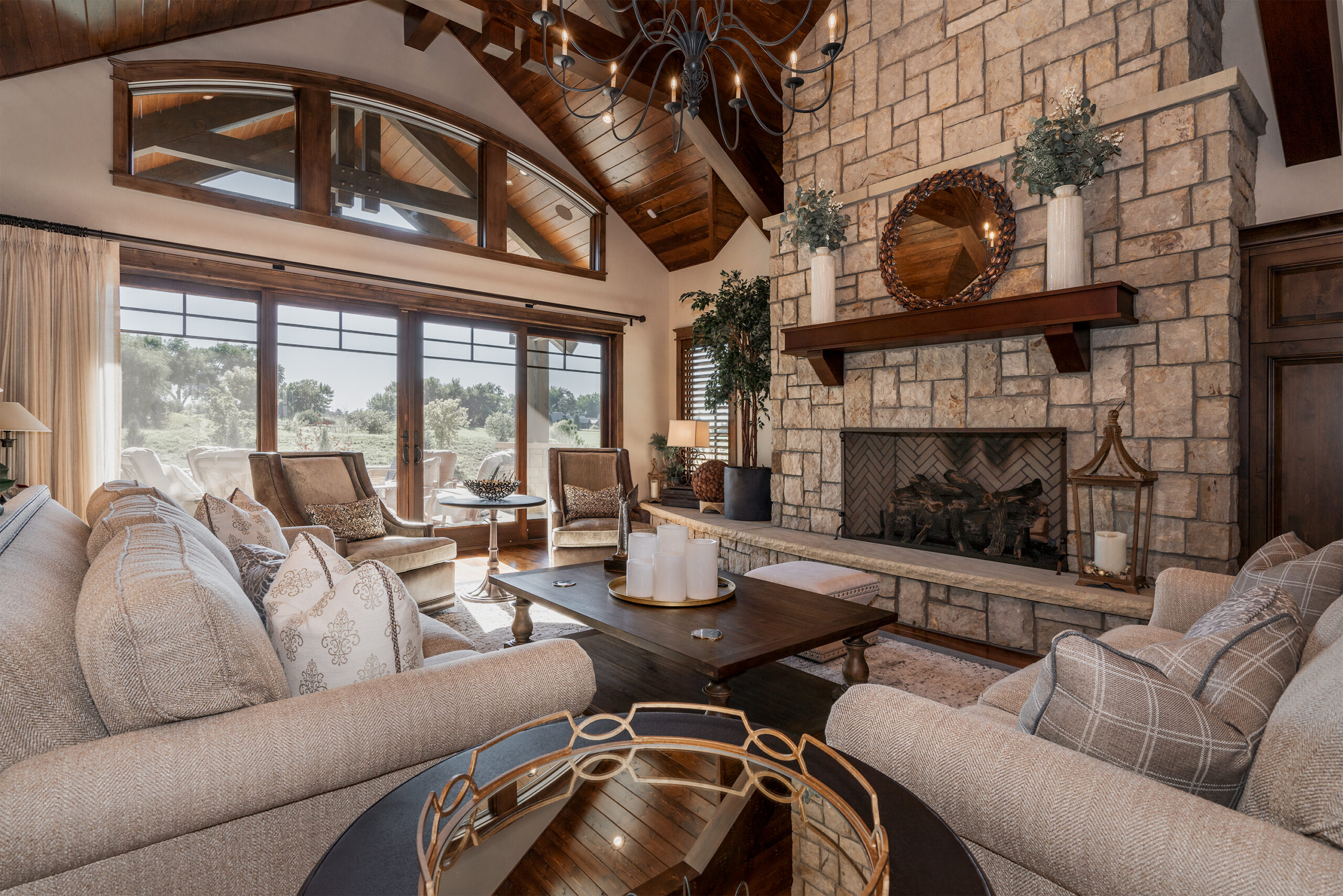 A bright living room with large floor to ceiling windows, stone wall fireplace, and stuffed sand colored chairs and sofa.