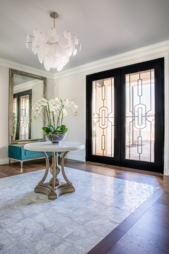 The entryway of a home with a chandelier, large glass doors, intricate floor tile pattern, and a table with a large orchid on top.
