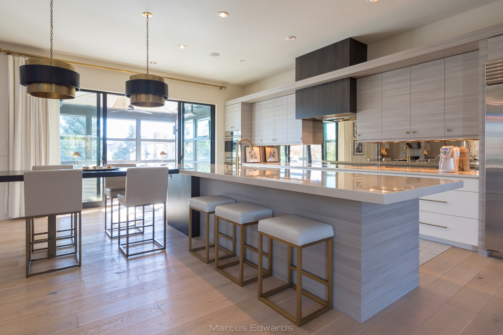 A bright kitchen with white marble counters, stools underneath, floor to ceiling windows, and white cabinets.