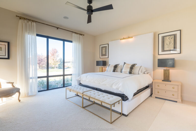 A bedroom with a large white bed, side table, chair, and floor to ceiling windows.