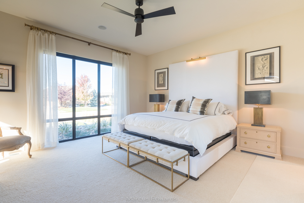 A bedroom with a large white bed, side table, chair, and floor to ceiling windows.