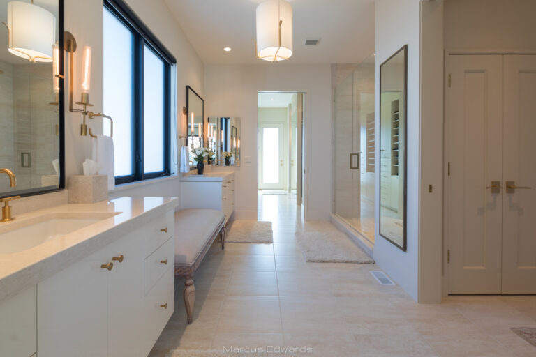 A primary bathroom with white fixtures, hanging lights, mirrors, and bright windows.