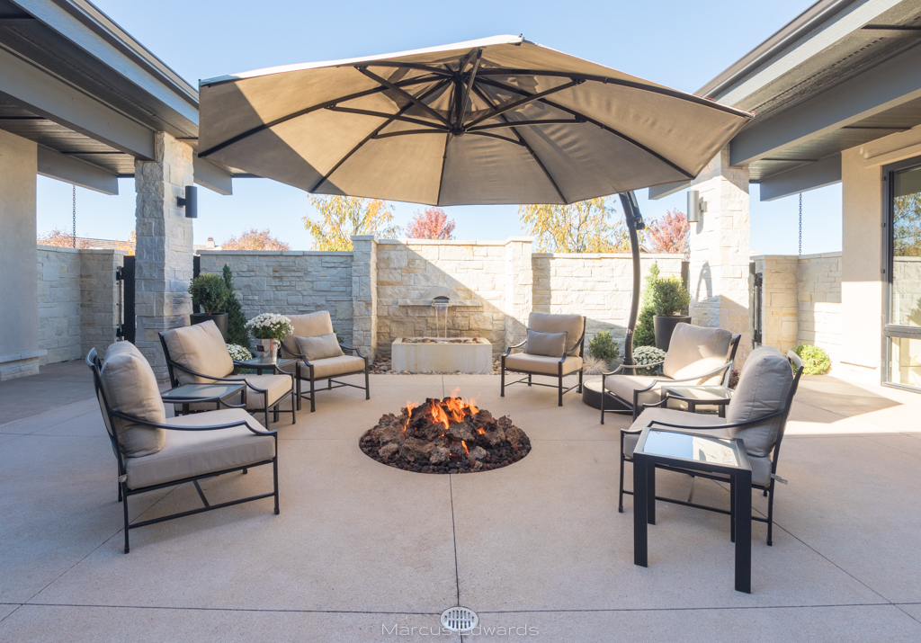 A stone walled patio space with a built in electric fireplace in the ground, large covered umbrella, and comfy chairs.